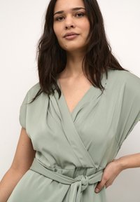 Light green wrap dress with short sleeves, made from a smooth material, featuring a tie belt and pleated design accenting the neckline.