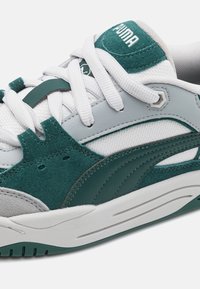 Tenisky Puma s horní částí z zelené semišové kůže a šedé kůže, bílými síťovinovými detaily a plochými bílými tkaničkami. Silná bílá gumová podrážka.