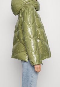 Vue latérale d'une personne portant une doudoune brillante vert olive avec une grande capuche et un jean bleu clair sur un fond uni.