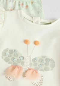 Wit T-shirt met korte mouwen met een vlinderontwerp, groene gestippelde vleugels, lichte roze tule accenten en oranje pompondetails.
