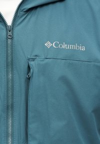 Τζάκετ σε χρώμα πετρόλ με φερμουάρ, με ορατό φερμουάρ και θήκη στο στήθος με φερμουάρ, με την επωνυμία και το λογότυπο "Columbia" στην αριστερή πλευρά.