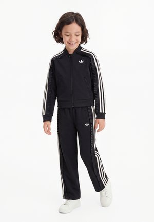 Kind lacht en kijkt naar beneden, draagt een zwarte Adidas trainingspak met witte strepen en witte sneakers tegen een effen witte achtergrond.