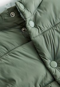 Detail einer grünen Pufferjacke mit gestepptem Design, Druckknöpfen und einer glatten, glänzenden Materialoberfläche. Sichtbare enge Nahtnähte.