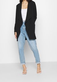 Giacca nera drappeggiata con apertura frontale, abbinata a jeans a vita alta di un blu chiaro. Il modello indossa sandali bianchi con cinturini. Tessuto liscio e leggero.