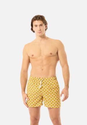 Homme torse nu debout, portant un short de bain jaune avec un motif géométrique circulaire blanc et un cordon de serrage blanc sur un fond clair.