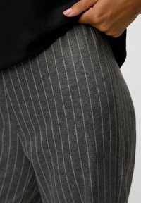 Une main tire un t-shirt noir, révélant des leggings gris avec de fines rayures verticales blanches sur la hanche et le haut de la cuisse de la personne.