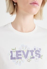 Biely bavlnený tričko s logom "LEVI'S" v purpurovej farbe, obklopené kvetinovými dizajnmi v modrej, zelenej a žltej farbe. Okrouhly výstrih, krátke rukávy.