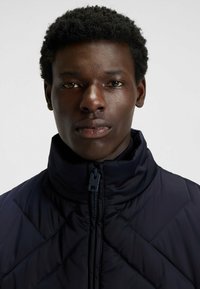 Mörkblå vadderad jacka med hög krage, quiltad textur och framdragkedja. Har diagonala sömmar och liten logotyp på dragkedjan.
