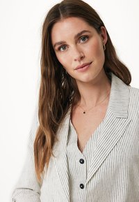 Kremowa koszula w pionowe czarne paski, z kołnierzykiem i czarnymi guzikami. Modelka nosi delikatny złoty naszyjnik i kolczyki w kształcie kółek.