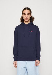 Marineblaues Baumwoll-Hoodie mit Kängurutasche, das ein kleines Logo in Rot, Weiß und Blau auf der linken Brust trägt. Mit verstellbarem Kapuzenzug.
