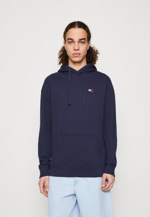 Sudadera de algodón azul marino con un bolsillo tipo canguro, que presenta un pequeño logotipo en rojo, blanco y azul en el lado izquierdo del pecho. Capucha con cordón.