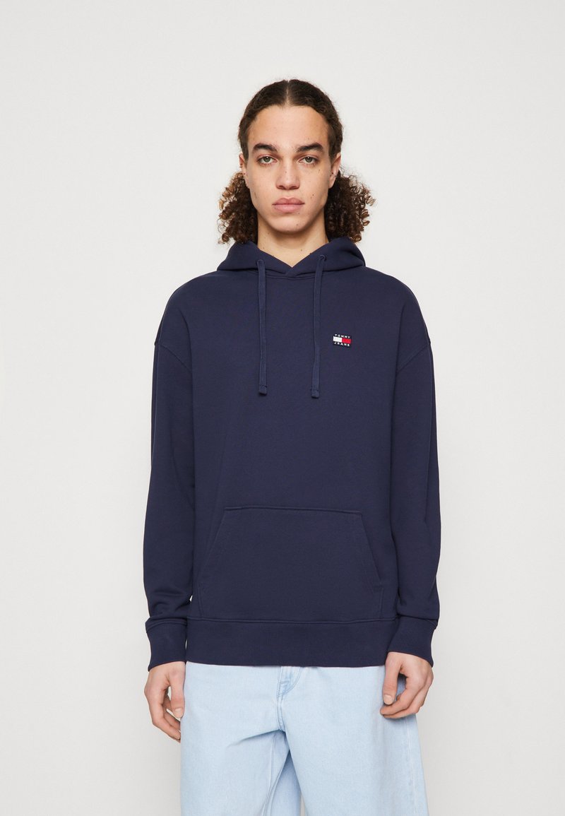 Marineblaues Baumwoll-Hoodie mit Kängurutasche, das ein kleines Logo in Rot, Weiß und Blau auf der linken Brust trägt. Mit verstellbarem Kapuzenzug.