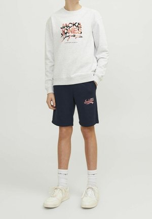 Grauer Pullover mit Logo auf der Vorderseite, kombiniert mit navyblauen Shorts mit rotem Logo. Weiße Sneaker und sichtbare weiße Socken vervollständigen das Outfit.