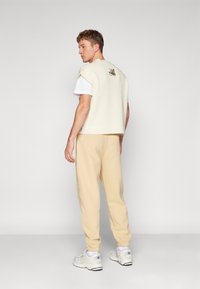 Roomkleurige gebreide vest met korte mouwen, gedragen over een witte shirt, gecombineerd met beige sweatpants en witte sneakers. Heeft een geborduurd detail.