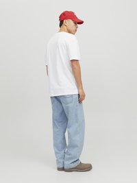 Weißes T-Shirt, hellblaue lässige Jeans und braune Schuhe. Zubehör umfasst eine rot rückwärts getragene Kappe. Einfaches Design, lässige Passform.