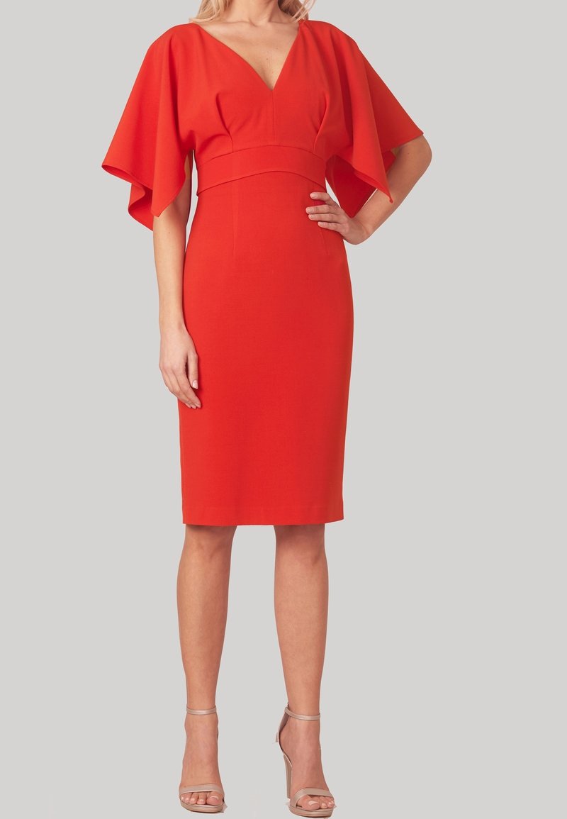 Femme portant une robe rouge ajustée longueur genou avec de larges manches courtes fluides et des sandales à talons hauts couleur chair, debout devant un fond uni.