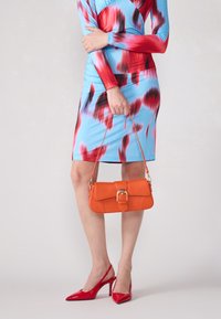 Sac à main en cuir orange de forme rectangulaire, avec un détail de boucle et une sangle détachable. Associé à une robe à motifs multicolores et des talons rouges.