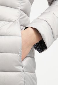 Ręka wsunięta do zamkniętej kieszeni jasnoszarej pikowanej kurtki, w której widoczna jest podszewka rękawa w kropki.