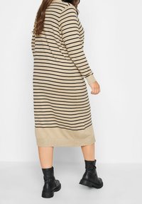 Béžové žebrované pletené šaty s černými vodorovnými pruhy, dlouhými rukávy a přímým střihem, doplněné černými kotníkovými botami.