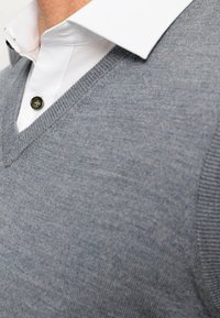 Grijze gebreide trui met een V-hals, met een fijne textuur. Gepaard met een witte blouse met kraag, met zwarte knopen op de kraag.