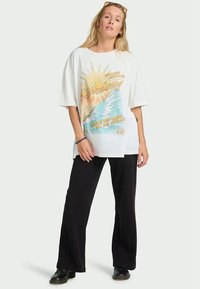 Λευκό oversize t-shirt με πολύχρωμο σχέδιο ήλιου και κύματος. Συνδυασμένο με μαύρες φαρδιές παντελόνες και μαύρες μπότες.