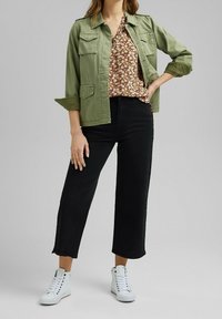Giacca verde oliva con due tasche frontali, blusa floreale, jeans neri a gamba larga e sneakers bianche alta. Texture liscia, stile casual.