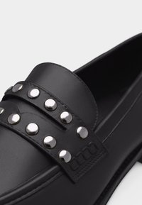 Mocassin en cuir noir avec deux sangles ornées de clous ronds argentés sur le dessus, présenté en gros plan détaillé.