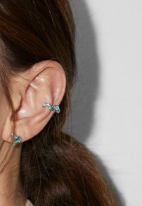 Oreja con dos pendientes de plata que presentan pequeñas piedras preciosas azules y verdes, sobre fondo de cabello castaño y un fondo claro sencillo.