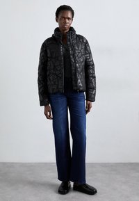Zwarte gewatteerde pufferjack met een capuchon, voorzien van abstracte grijze patronen, gecombineerd met donkere jeans en zwarte instapchoenen.