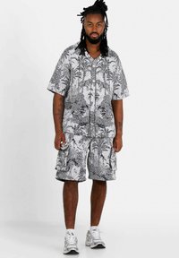 Camicia a maniche corte con stampa tropicale bianco e nero abbinata a pantaloni cargo coordinati; presenta motivi dettagliati e bottoni, indossata con sneakers bianche.