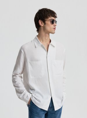 Giovane uomo che indossa occhiali da sole, una camicia bianca con due tasche sul petto e jeans blu, in piedi con le mani nelle tasche, rivolto verso destra.