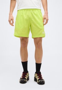 Άτομο που φοράει φωτεινά λαχανί σορτς, μαύρες κάλτσες μέχρι τη μέση της γάμπας και μαύρα και μπεζ παπούτσια για τρέξιμο σε μονοπάτι με κόκκινα κορδόνια.