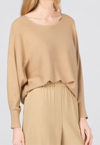 Beżowy sweter dzianinowy o szerokim dekolcie, opadających ramionach i długich rękawach, zestawiony z beżowymi spodniami z elastycznym pasem o miękkiej teksturze.