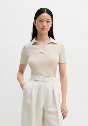 Vrouw met lang zwart haar gekleed in een beige top met korte mouwen en kraag, en een hooggetailleerde geplooide broek in off-white, staand met één hand in de zak.