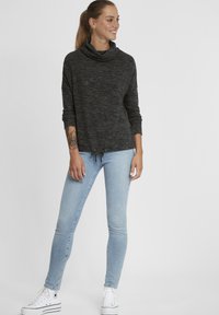 Maglione a collo alto grigio scuro con maniche lunghe, realizzato in un tessuto lavorato a maglia. Indossato con jeans skinny azzurri e sneaker bianche. Dettaglio con cordoncino sull'orlo.