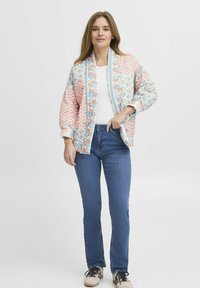 Giacca trapuntata in colori pastello con motivi floreali e geometrici, dotata di colletto e polsini a coste, abbinata a jeans blu e sneakers.