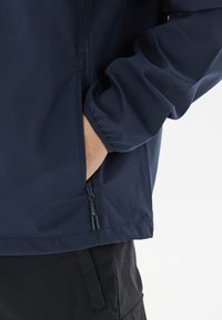 Mörkblå jacka med en slät yta, utrustad med en dragkedjeficka och elastiska ärmslut, designad för komfort och funktionalitet.