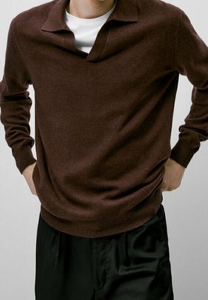 Mężczyzna ubrany w ciemnobrązowy sweter z długim rękawem na białą koszulę, w luźnych czarnych spodniach, z rękami opartymi na biodrach.