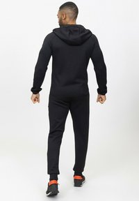 Haut de survêtement noir avec un motif texturé, associé à un bas de jogging assorti. Il présente une capuche et des accents orange contrastants sur les chaussures.