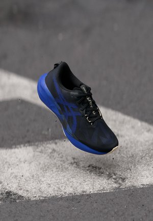 Chaussure de course noire et bleue avec lacets, en l'air au-dessus d'un bitume avec des marquages routiers blancs, petites particules de saleté visibles en dessous.