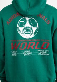 Green hooded sweatshirt featuring a soccer ball graphic, red text reading "FROM HERZOGENAURACH TO THE WORLD" with city names listed below.