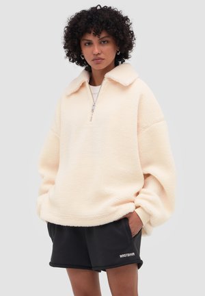 Femme aux cheveux bouclés portant un pull polaire crème avec une fermeture éclair et un short noir WRSTBHVR, les mains dans les poches.