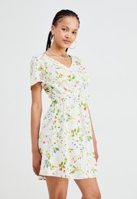 Witte jurk met bloemenprint, met een V-hals, korte mouwen en een striktaille. Versierd met groene en kleurrijke bloempatronen.
