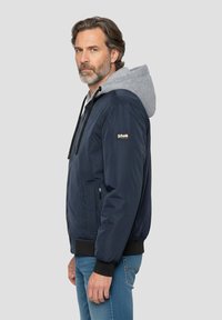 Marineblaue Bomberjacke mit grauem Kapuzenfutter, ausgestattet mit einem Reißverschluss, gerippten Bündchen und einem Logodetail am Ärmel. Blaue Jeans vervollständigen das Outfit.