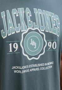 T-shirt in cotone verde scuro con testo bold bianco e azzurro chiaro, design del logo circolare e dettagli del marchio "JACK&JONES".