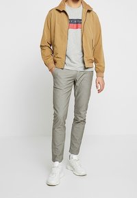 Ruskea pommitakki, harmaa t-paita, jossa on vaakasuora väriesto, vaaleanharmaat housut ja valkoiset tennarit. Rentoa asua, jossa on erilaisia tekstuureja.