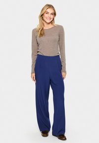 Lange mouwen beige top gecombineerd met marineblauwe wijde broek. De broek heeft een voorplooi, een relaxte pasvorm en een gladde stoftextuur. Bruine schoenen.