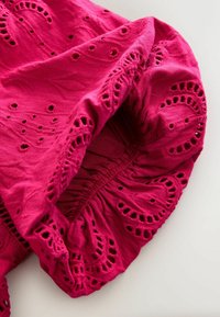Tissu rose vif avec broderie florale ajourée et bord élastique froncé, montrant la texture et les détails complexes de couture.