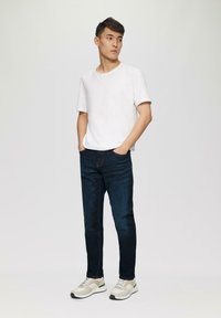 Weißes T-Shirt mit kurzen Ärmeln, dunkelblaue Jeans und hellfarbene Sneakers mit grauen und grünen Akzenten. Einfaches Design, figurbetonte Form.