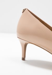 Chaussure à talon haut pour femme en cuir verni beige avec un talon fin de hauteur moyenne et un détail de couture lisse près du bord supérieur.
